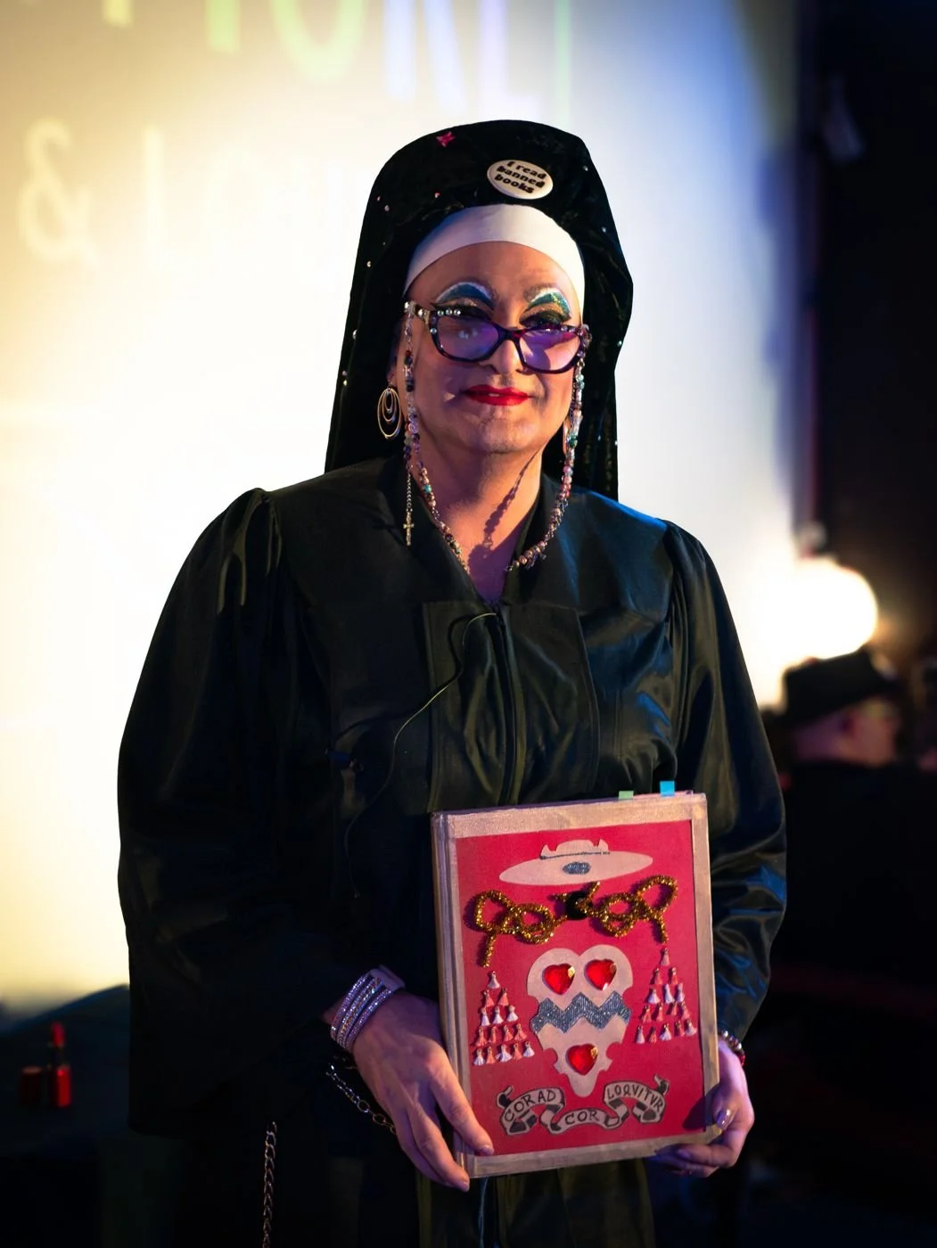 Sister Maggie in a dark robe and veil with round glasses, holding a decorated red book against her chest.