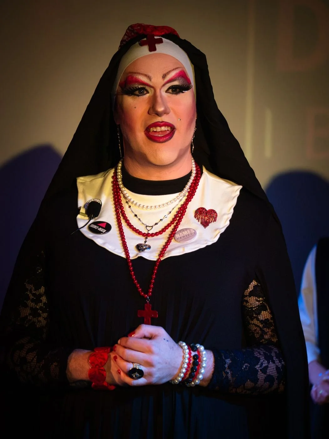 Sister Mercy in a black habit with a white collar, lace sleeves, and long beaded rosaries, holding a tube of lipstick.