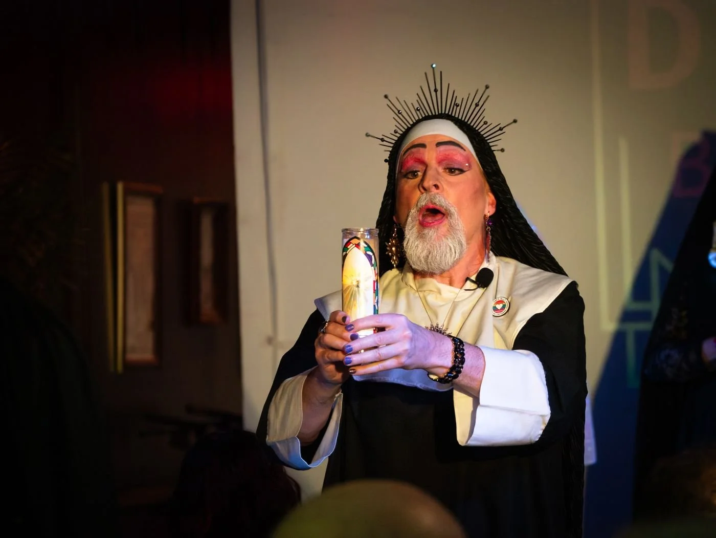 Sister Jude in a black habit with a white collar and a gray beard, holding a lit candle.