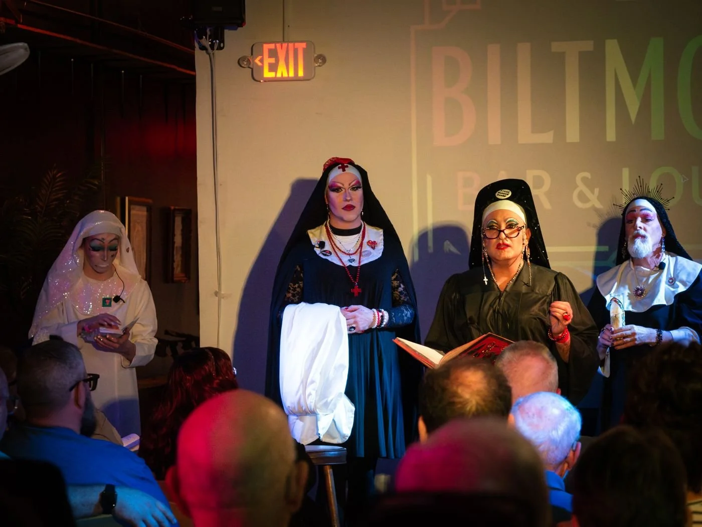 Sisters at the front of a bar room in front of a seated audience, one holding a white cloth, one reading from a book.