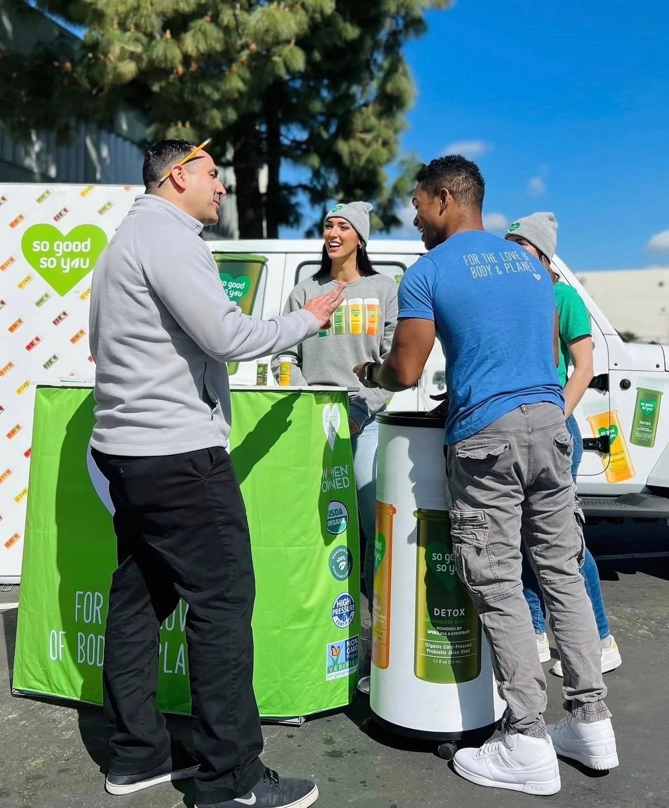 So Good So You ambassadors serving samples to adult shoppers at an outdoor sampling station, with a branded van behind