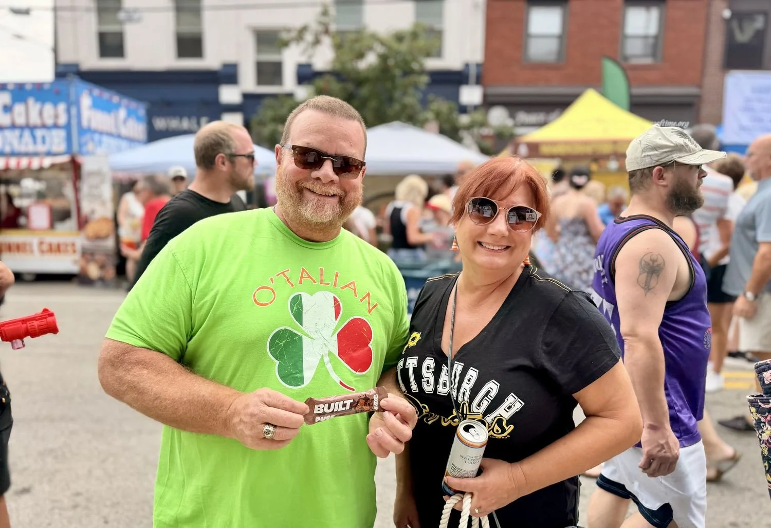 A smiling couple holding a Built protein bar and a drink at a street festival sampling stop