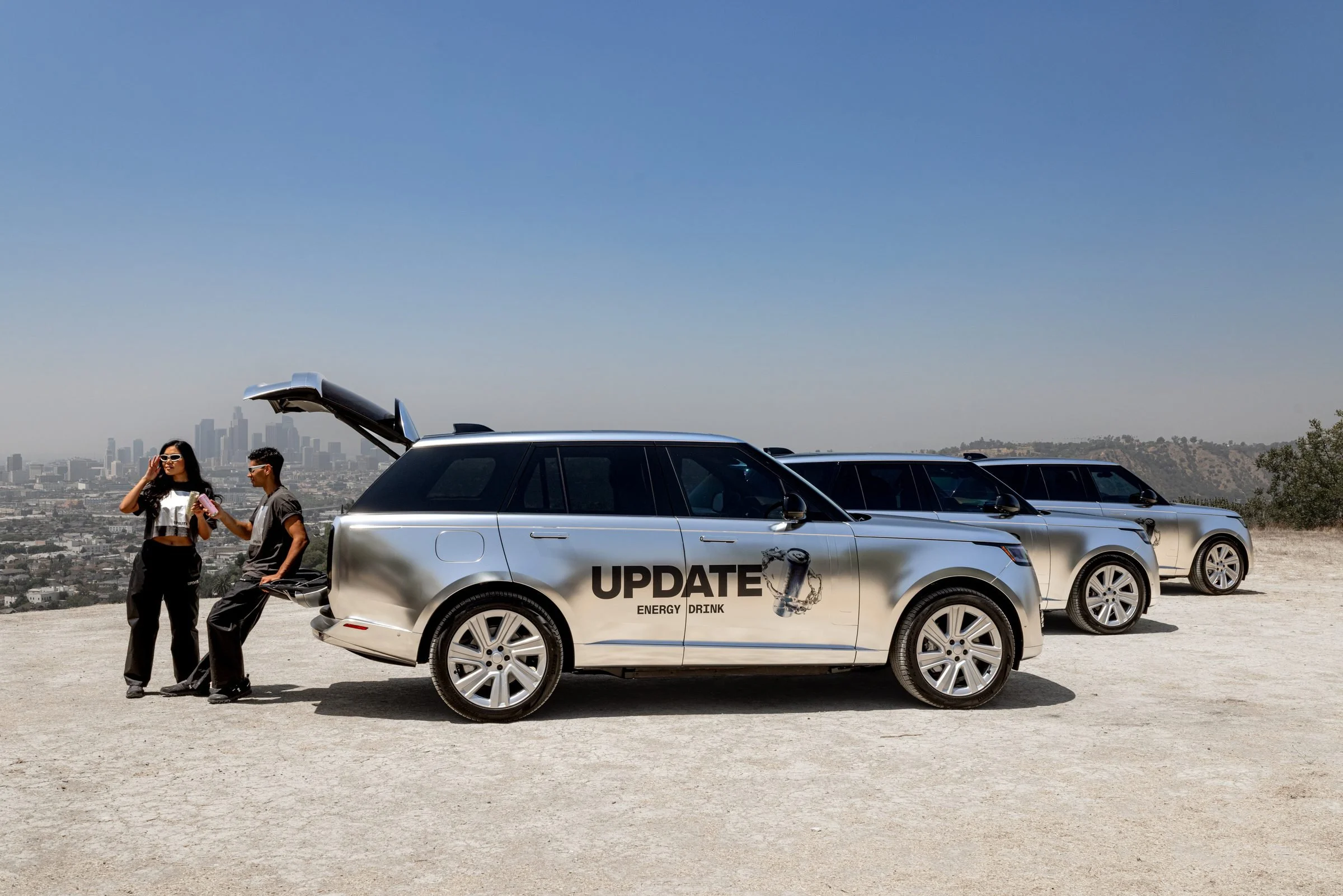 Two UPDATE Energy Drink ambassadors talking by the open tailgate of an UPDATE-wrapped Range Rover, with two more wrapped SUVs behind them