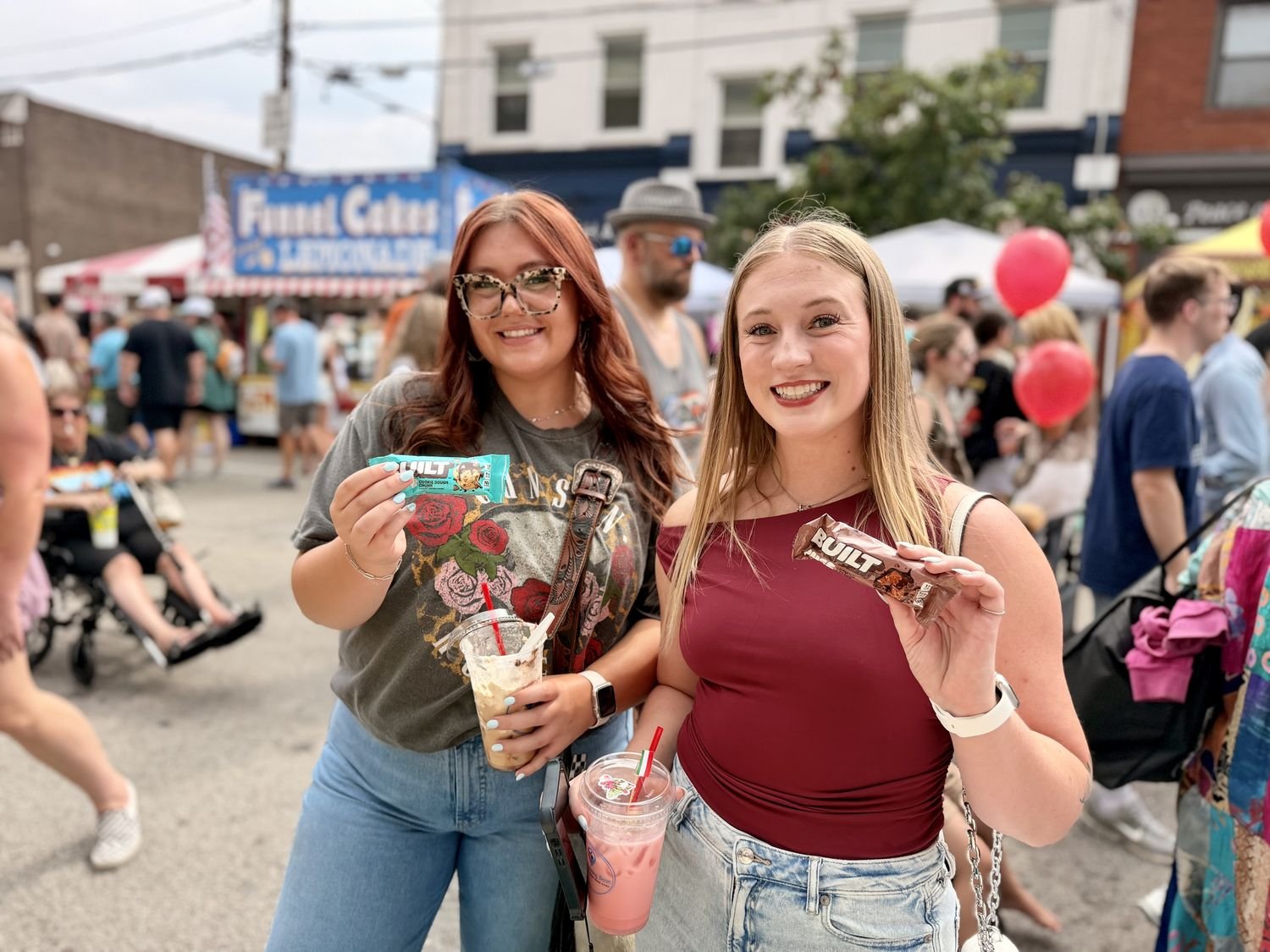 Smiling women holding Built protein bars and drinks at a festival event sampling stop