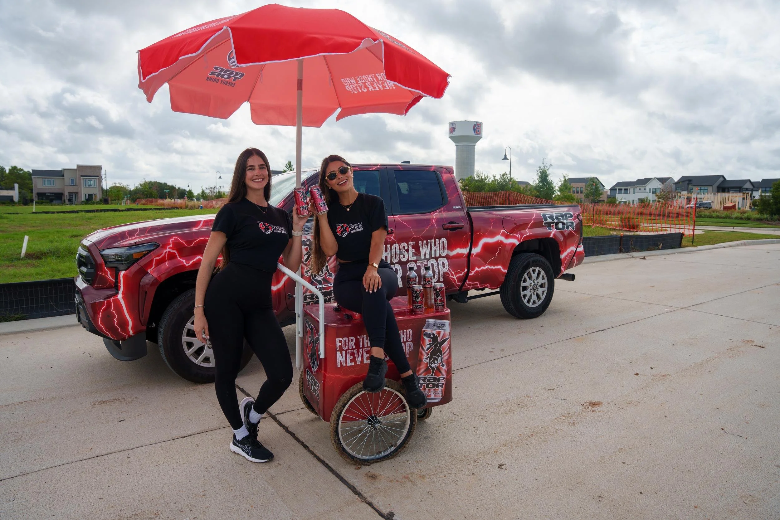 Two Raptor Energy ambassadors with a branded sampling cart and red umbrella in front of a fully wrapped Raptor Energy pickup truck