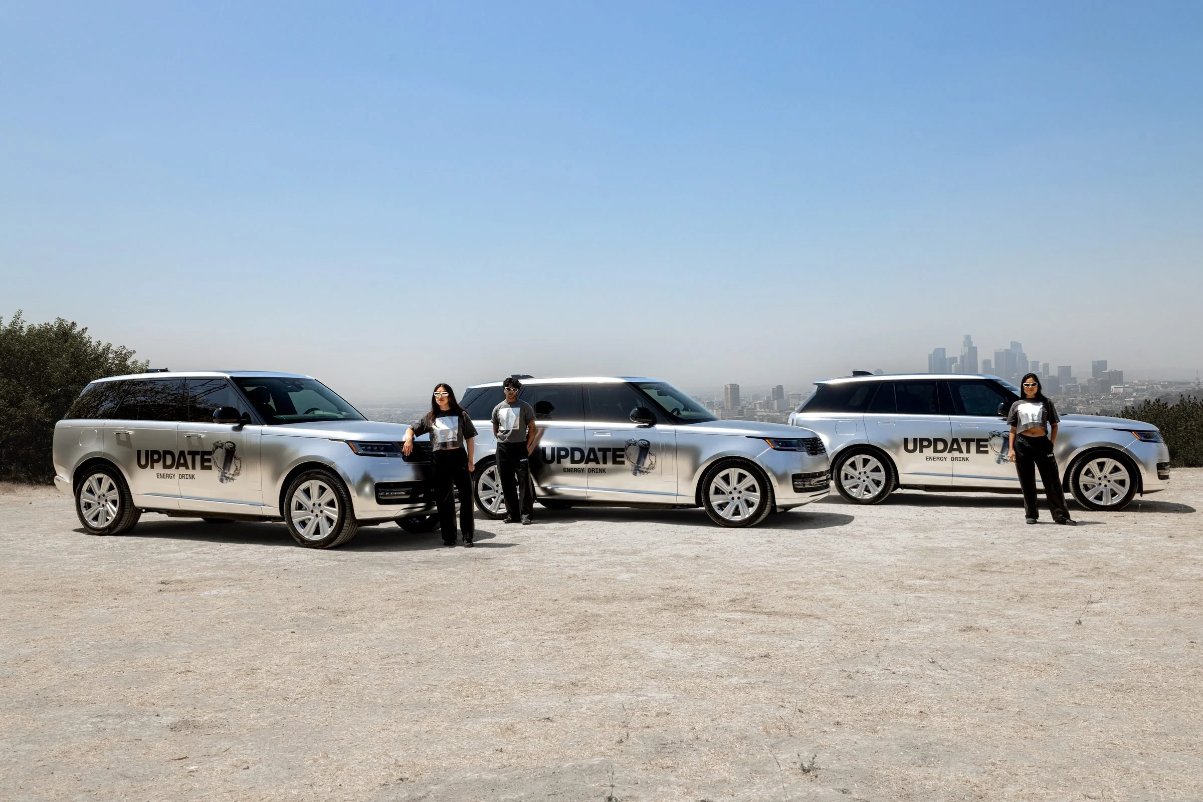 Branded UPDATE Range Rovers lined up with brand ambassadors beside them on a hilltop, downtown Los Angeles in the distance
