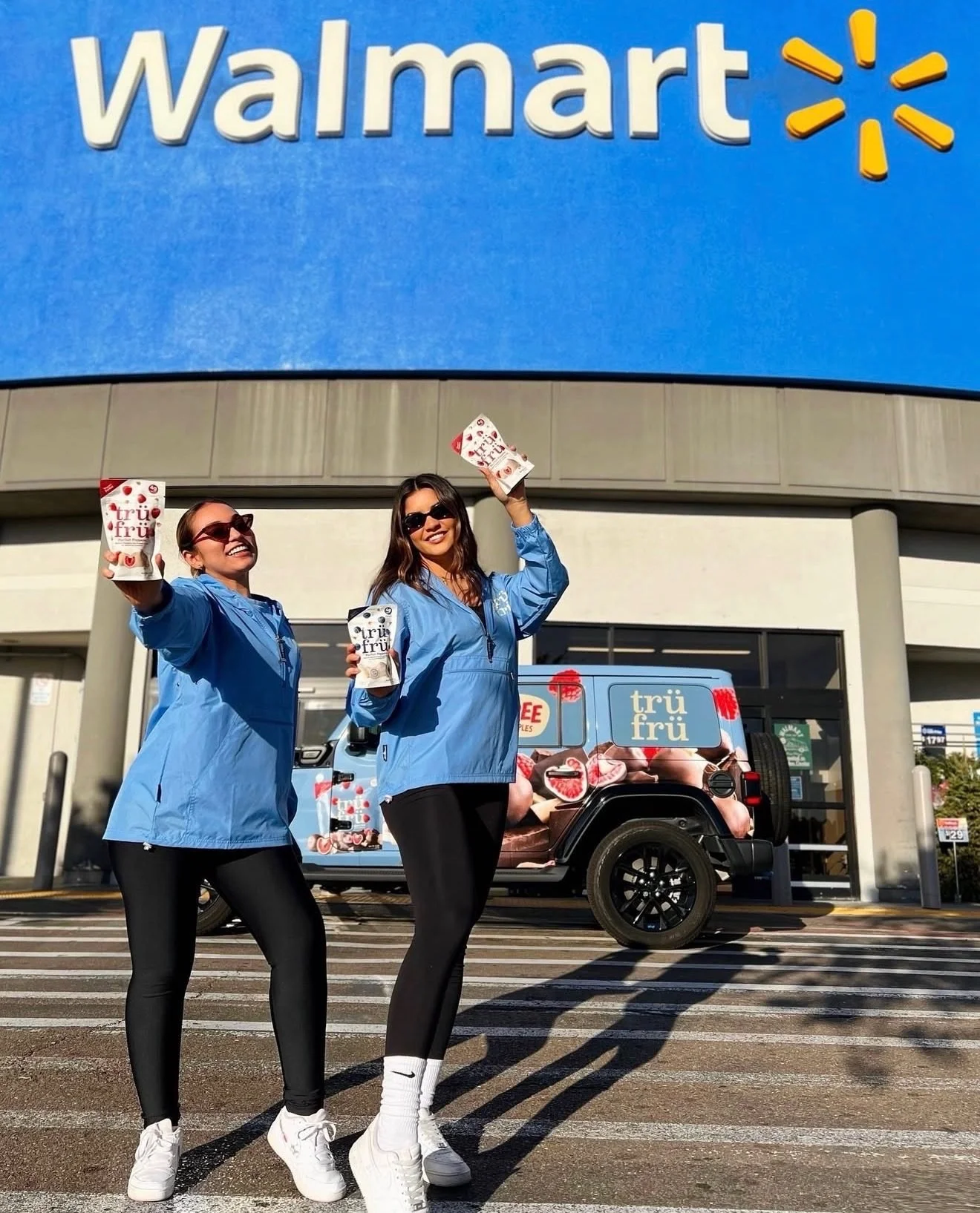 Two Tru Fru ambassadors cheering with product on a crosswalk under a Walmart sign, with a Tru Fru-wrapped Jeep behind them