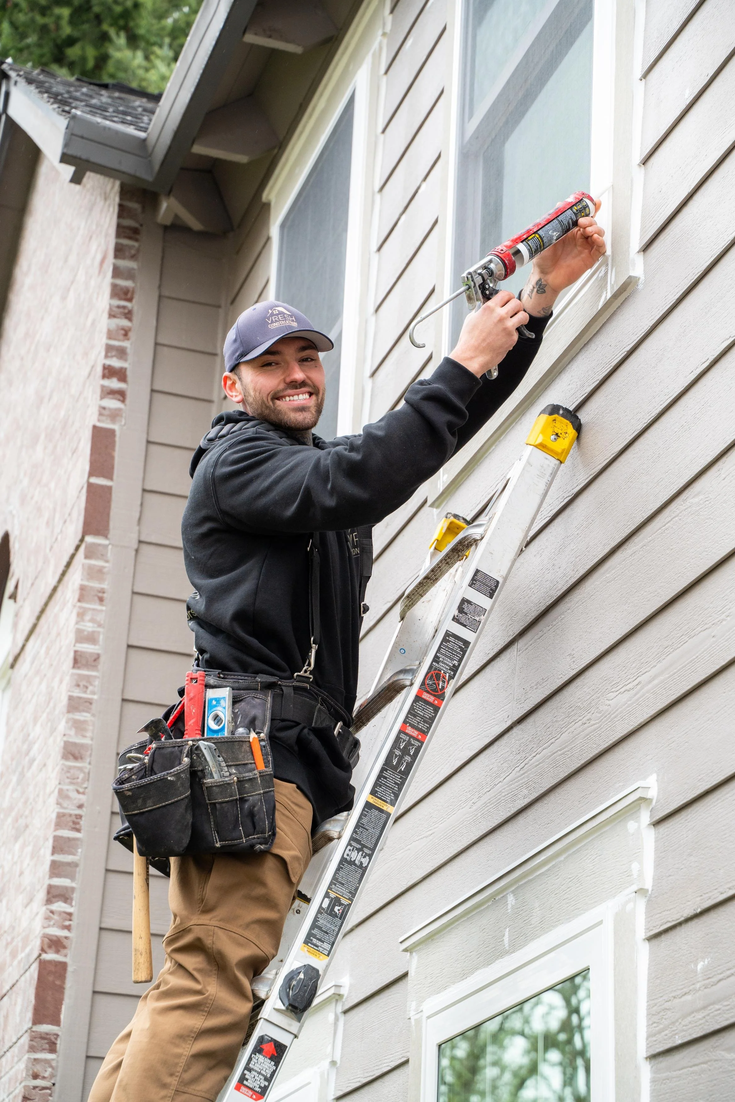 VResh technician on a ladder sealing around an upper window frame on a grey-sided Portland home