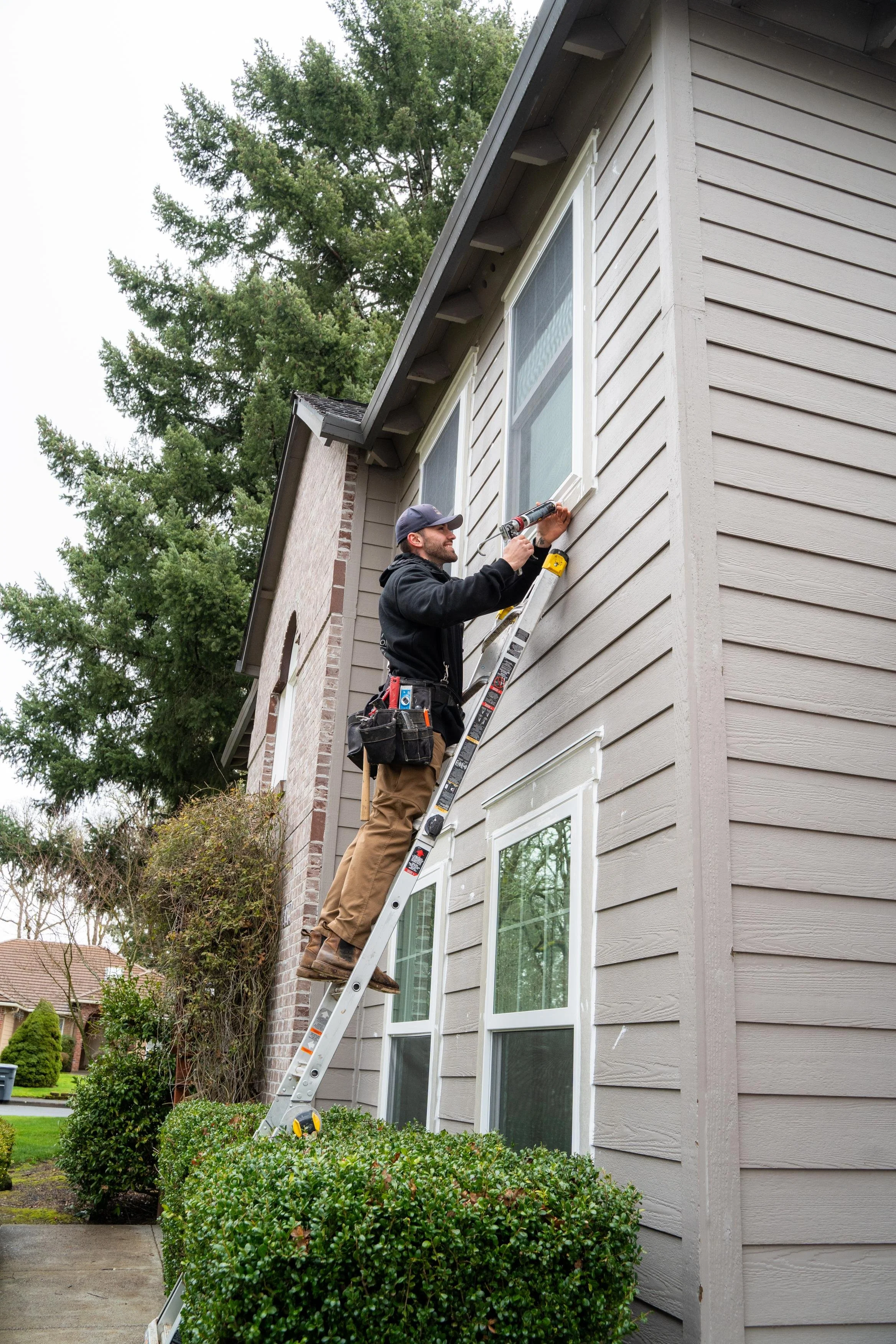 VResh Construction installer on a ladder caulking the trim of an upper window on a brick and siding home, for exterior door repair in Newberg, OR