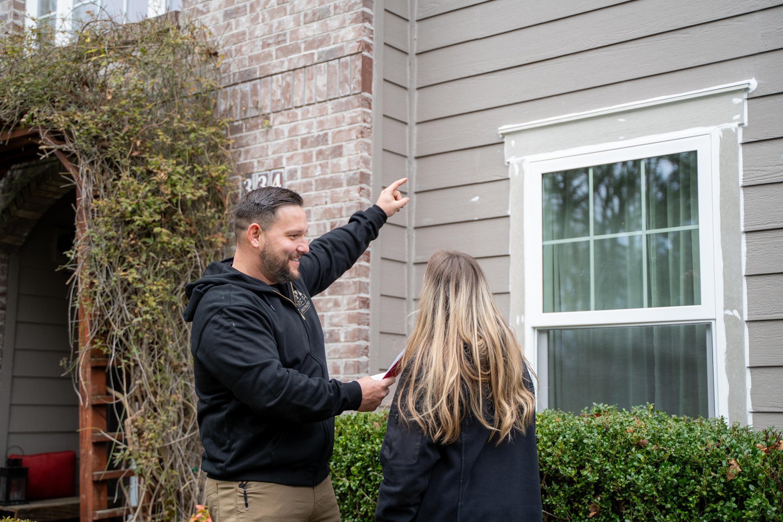 VResh owner pointing up at a white window on a grey-siding and brick home while a homeowner looks on