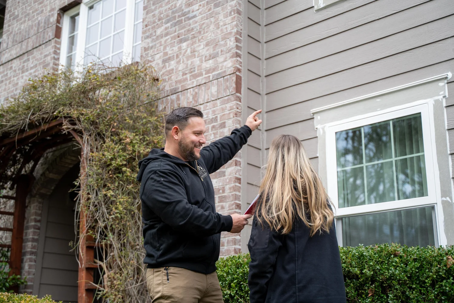 Vlad of VResh Construction pointing out the trim above a white window to a homeowner beside a brick and siding wall