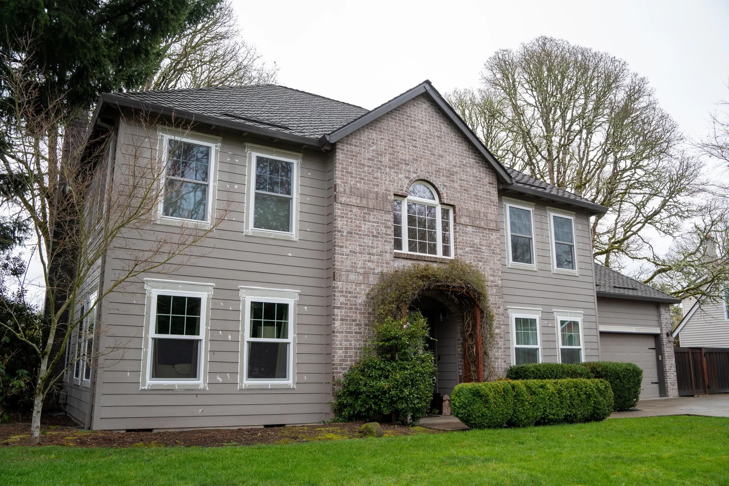 Two-storey grey brick and siding Portland home with an arched-top window in the brick gable above the vine-covered front entry, between rectangular windows