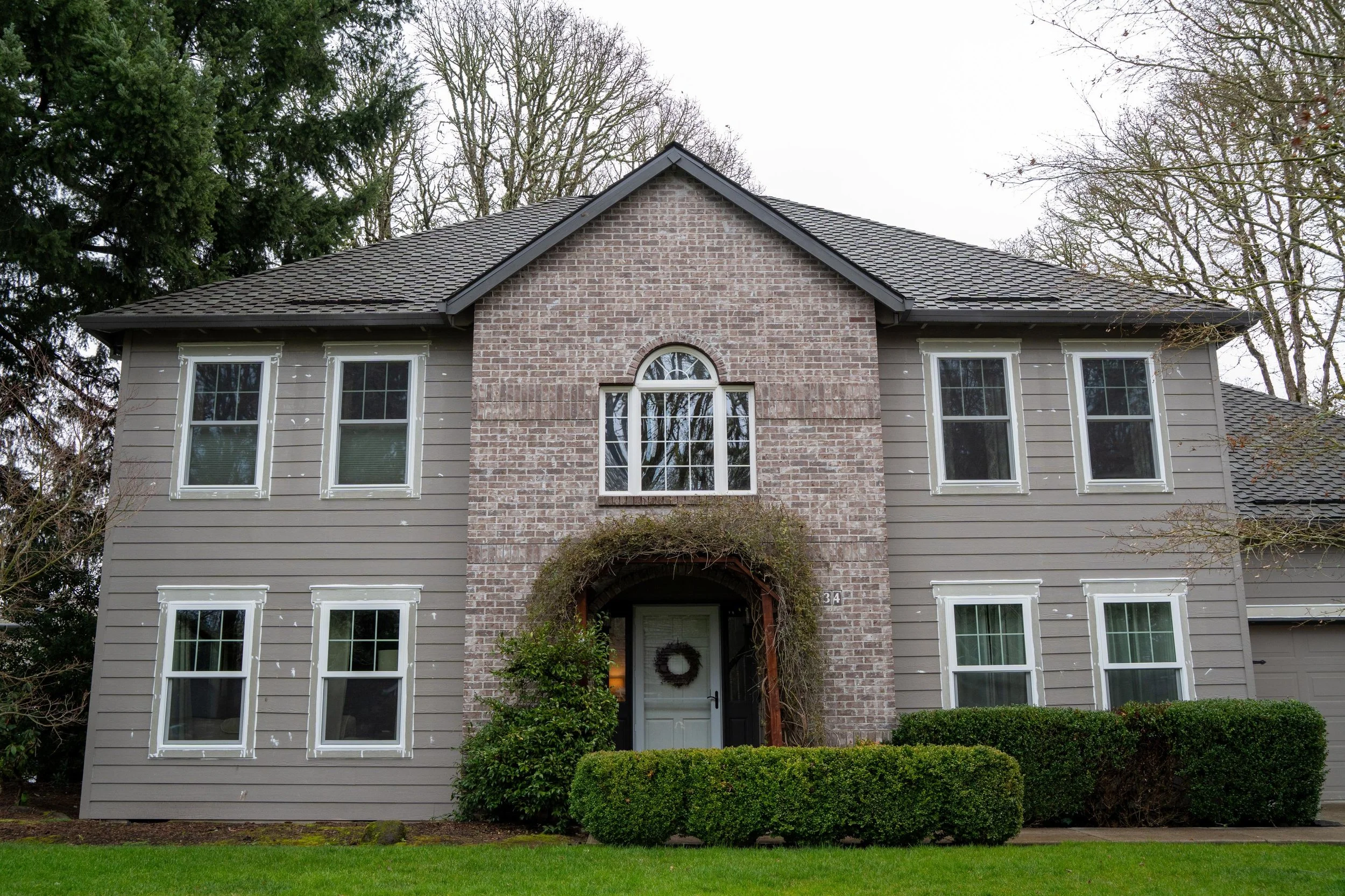 Front of a two-storey brick and siding home with white windows and a wreath on its front door under an arched trellis, for VResh Construction exterior door repair in Salem, OR