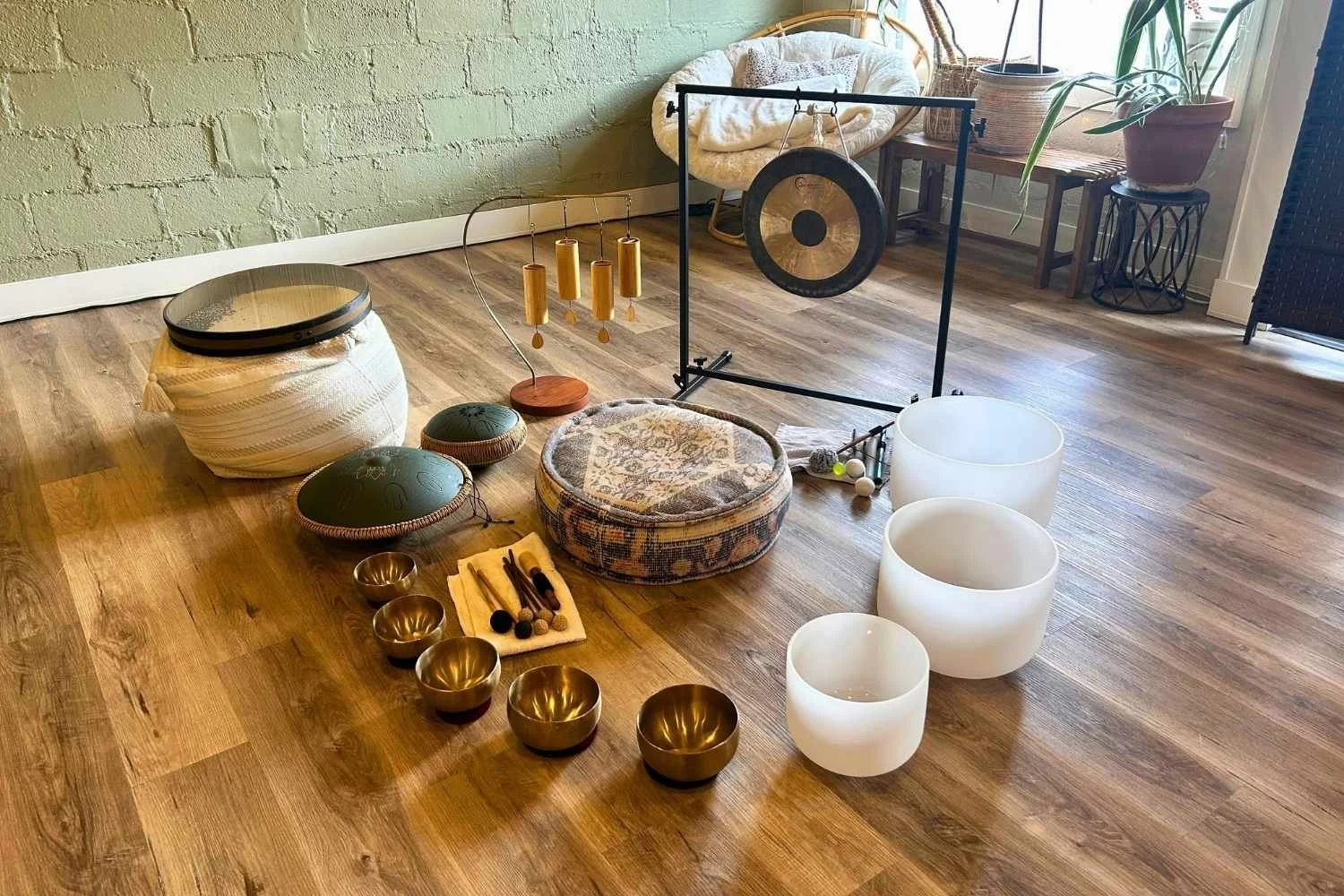 A gong on a stand beside crystal and bronze singing bowls, set up on a wooden floor before a sound bath.
