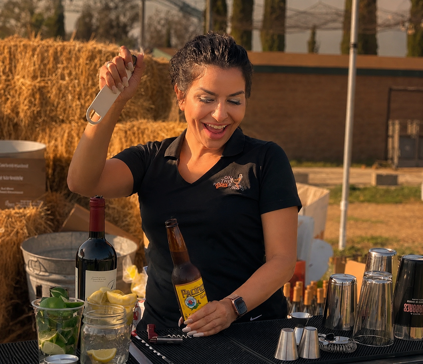 On The Rocks Girls bartender in a branded OTR polo opening a drink at a rustic outdoor Southern California event at golden hour