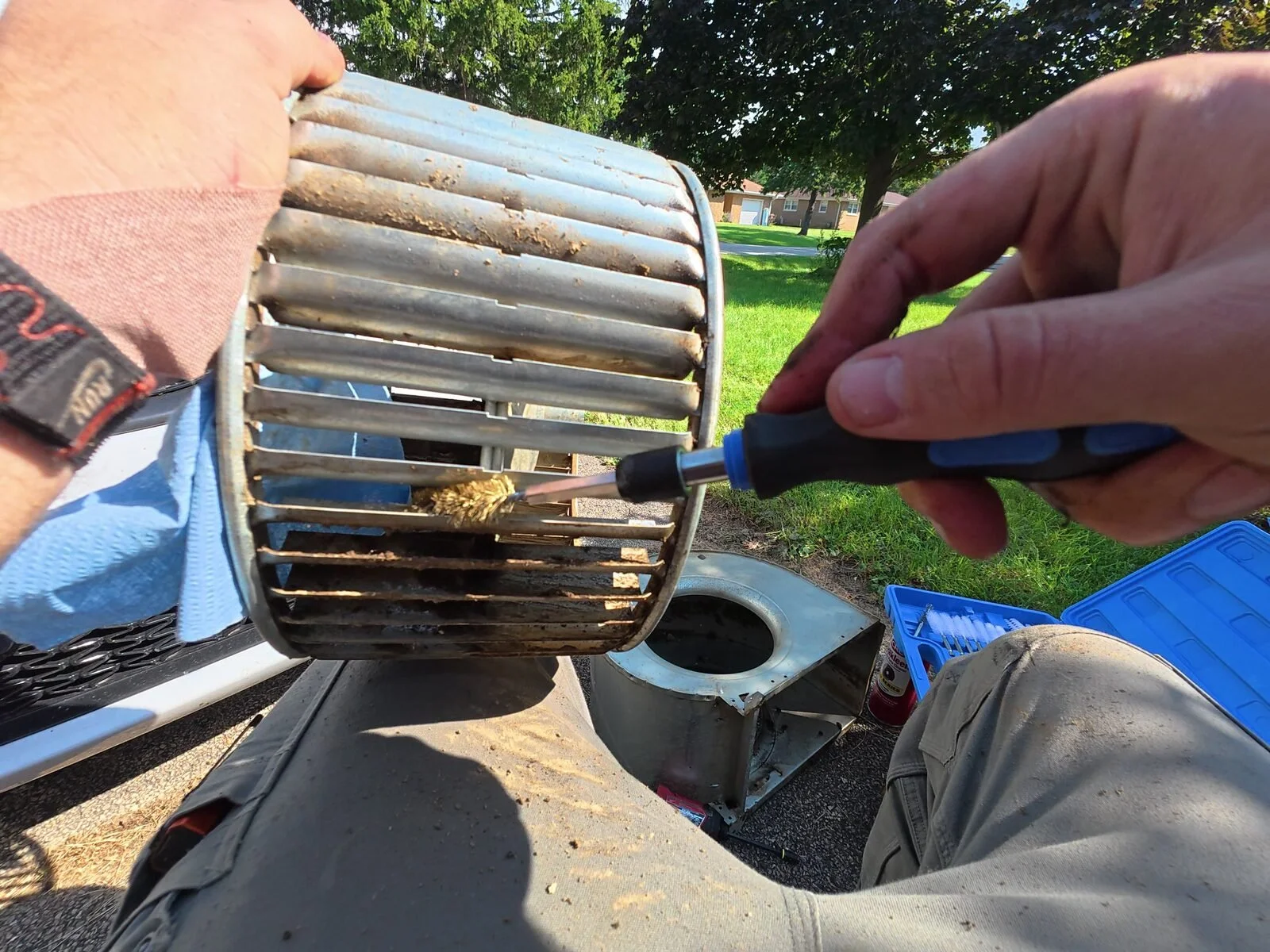 Generations HVAC technician using a hand brush to remove buildup from a blower wheel outdoors.