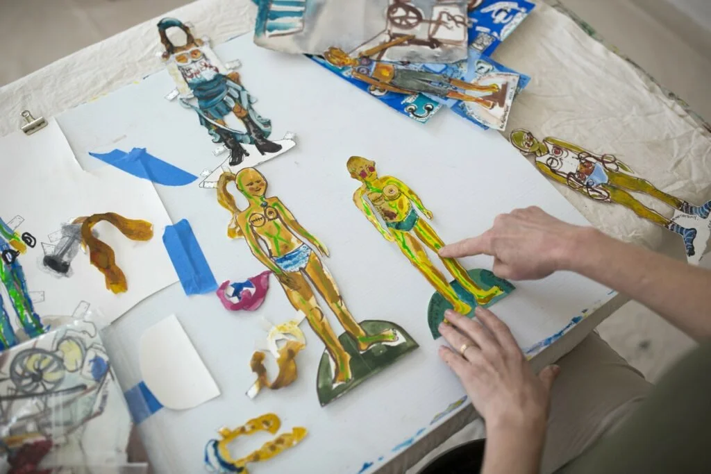 Painted paper dolls and interchangeable pieces arranged on Erin McGee Ferrell’s studio worktable.