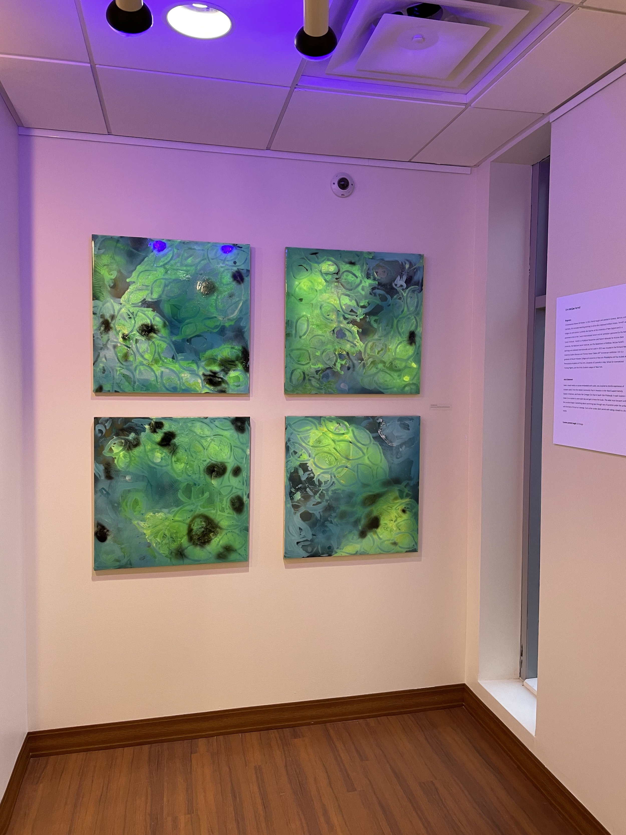 Four luminous green and blue paintings displayed under black lights at UNE Art Gallery in 2022.