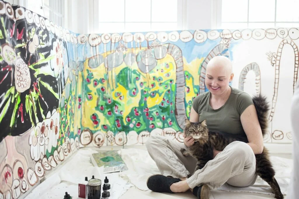Erin McGee Ferrell seated on her studio floor with her cat Milo, surrounded by paintings.