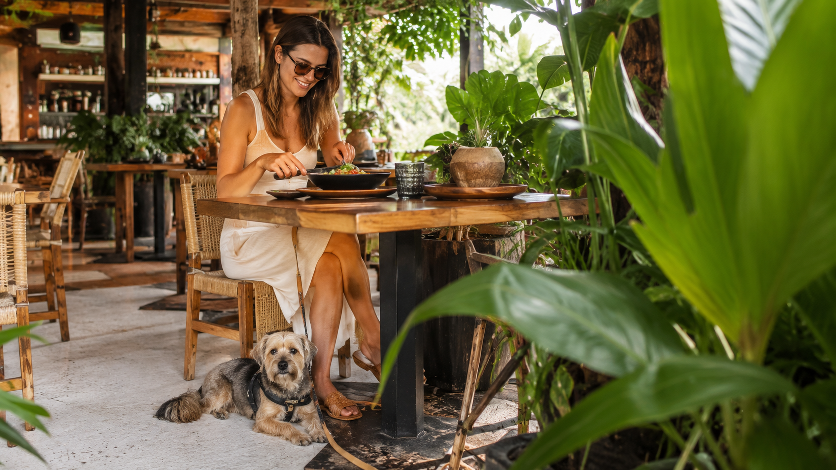 Woman enjoying lunch at an open-air jungle-inspired restaurant terrace in Playa del Carmen with a small dog resting under the table