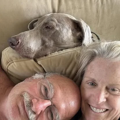 An older couple cuddling with their senior Weimaraner on the couch