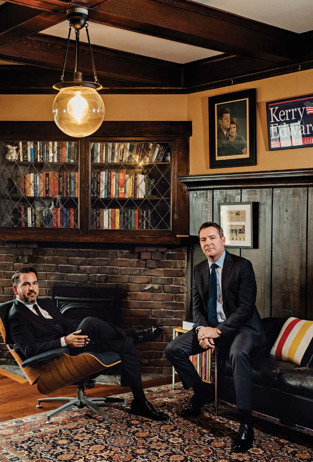 Nathan Ballard, seated in a leather lounge chair, and P.J. Johnston, seated on the arm of a sofa, in a wood-paneled office with glass-front bookcases and a brick fireplace.