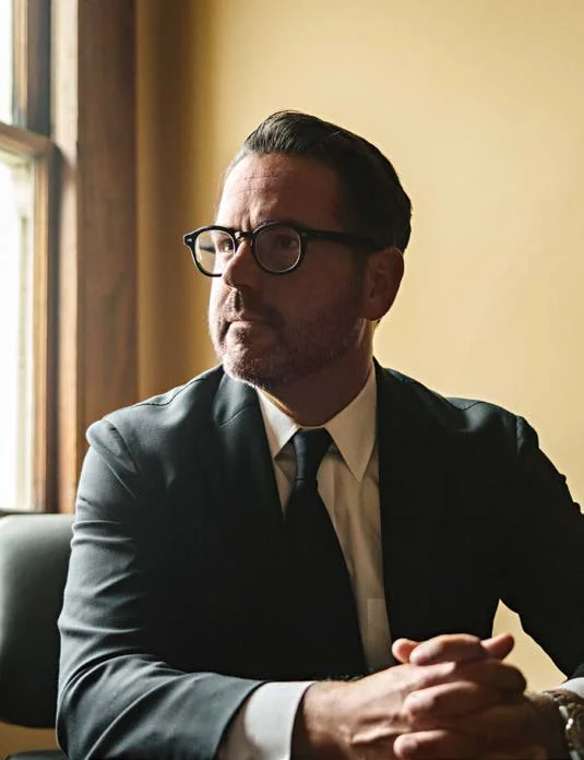 Nathan Ballard, in glasses and a dark suit, seated beside a window with his hands clasped.
