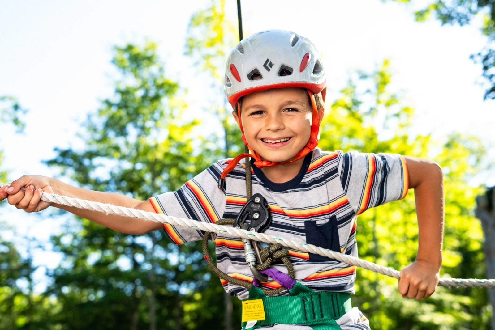 Victor smiling on the ropes course at Adventure Land NY — white and orange helmet, striped shirt