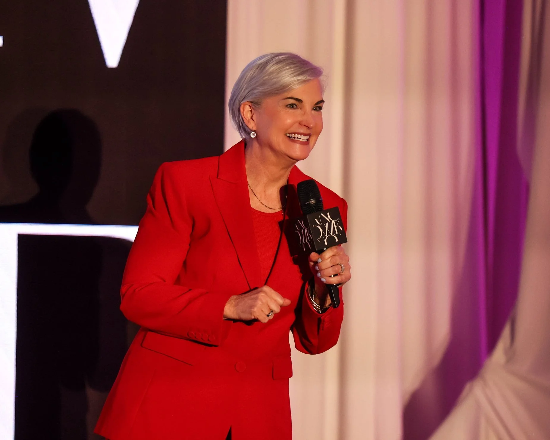 Christina Langdon speaking on stage in a red suit, microphone in hand, against the KNOW Summit screen