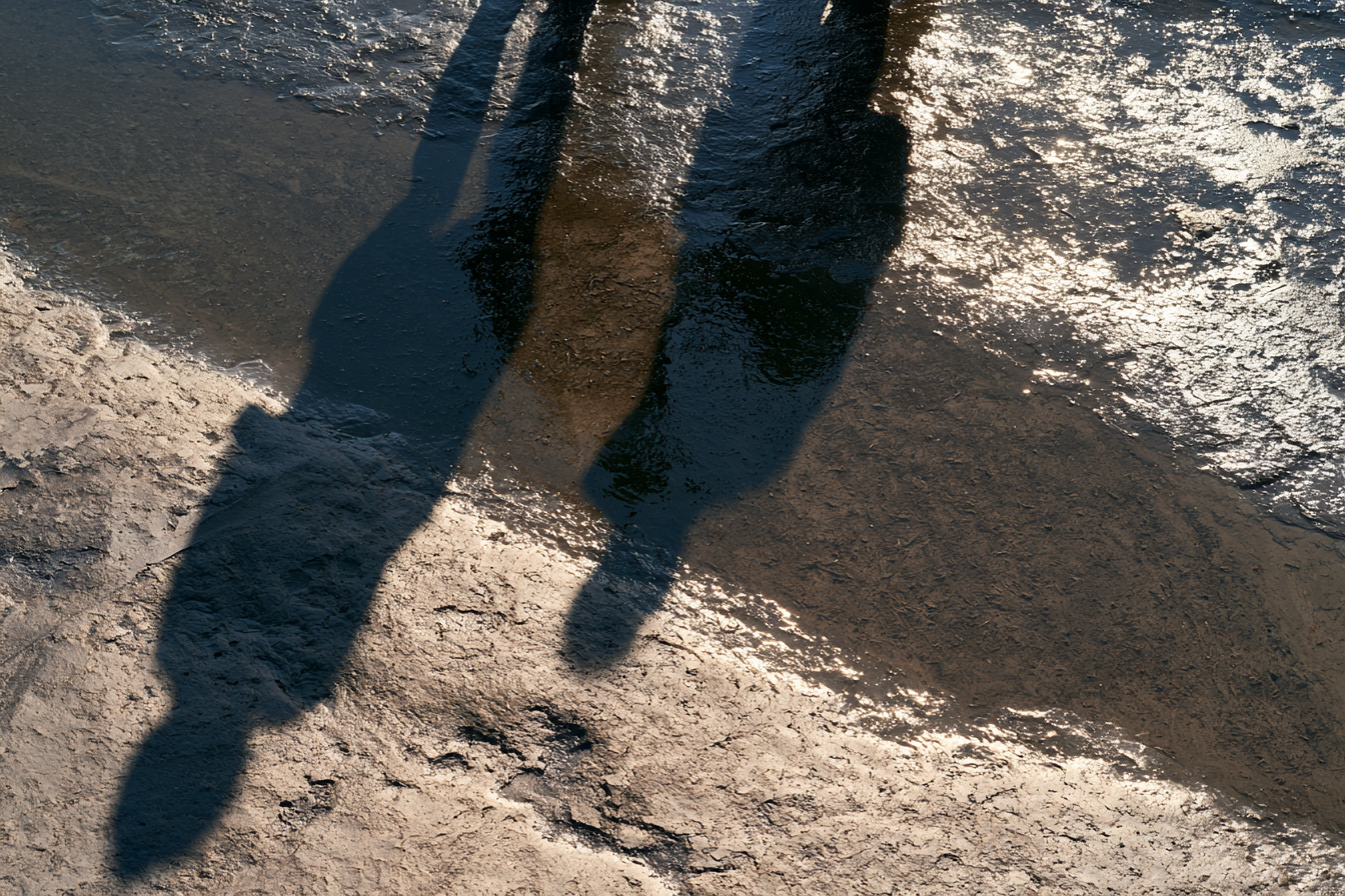Two elongated human-shaped shadows cross textured wet ground beside bright reflections.