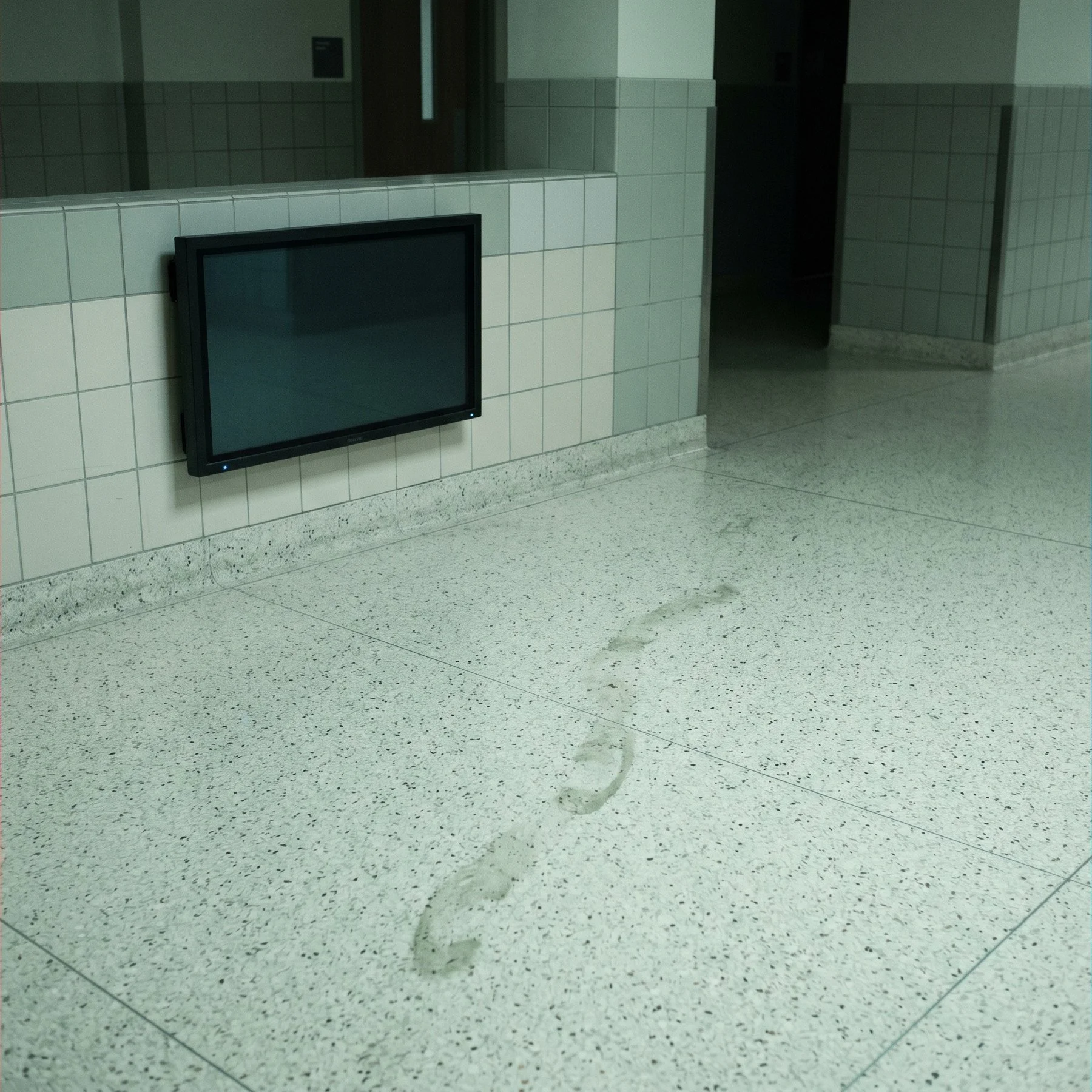 Faint wet footprints crossing a tiled floor beneath a dark wall-mounted screen.