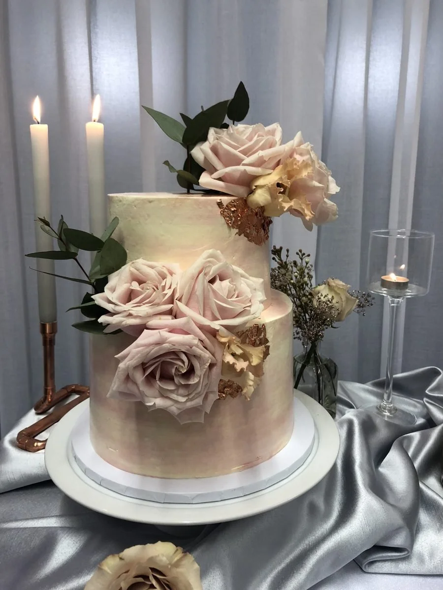 A two tier cake with blush roses and copper leaf beside tall candles