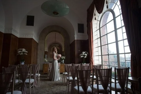 A bride standing among dressed chairs beside a tall arched window
