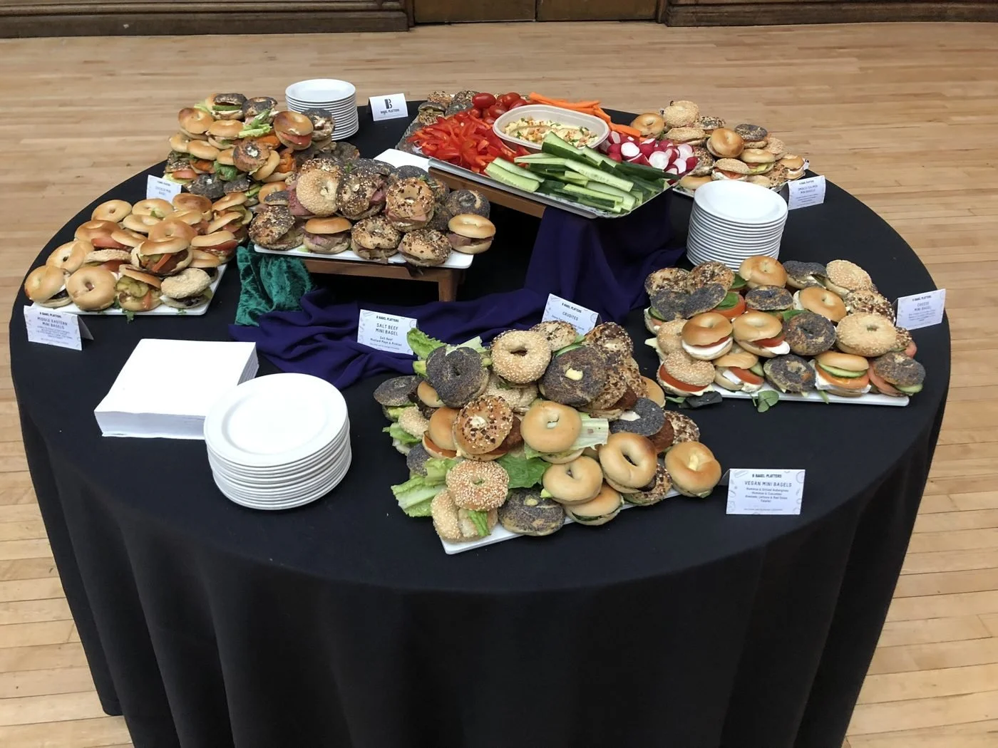 A round table of bagel platters and crudités dressed with velvet runners