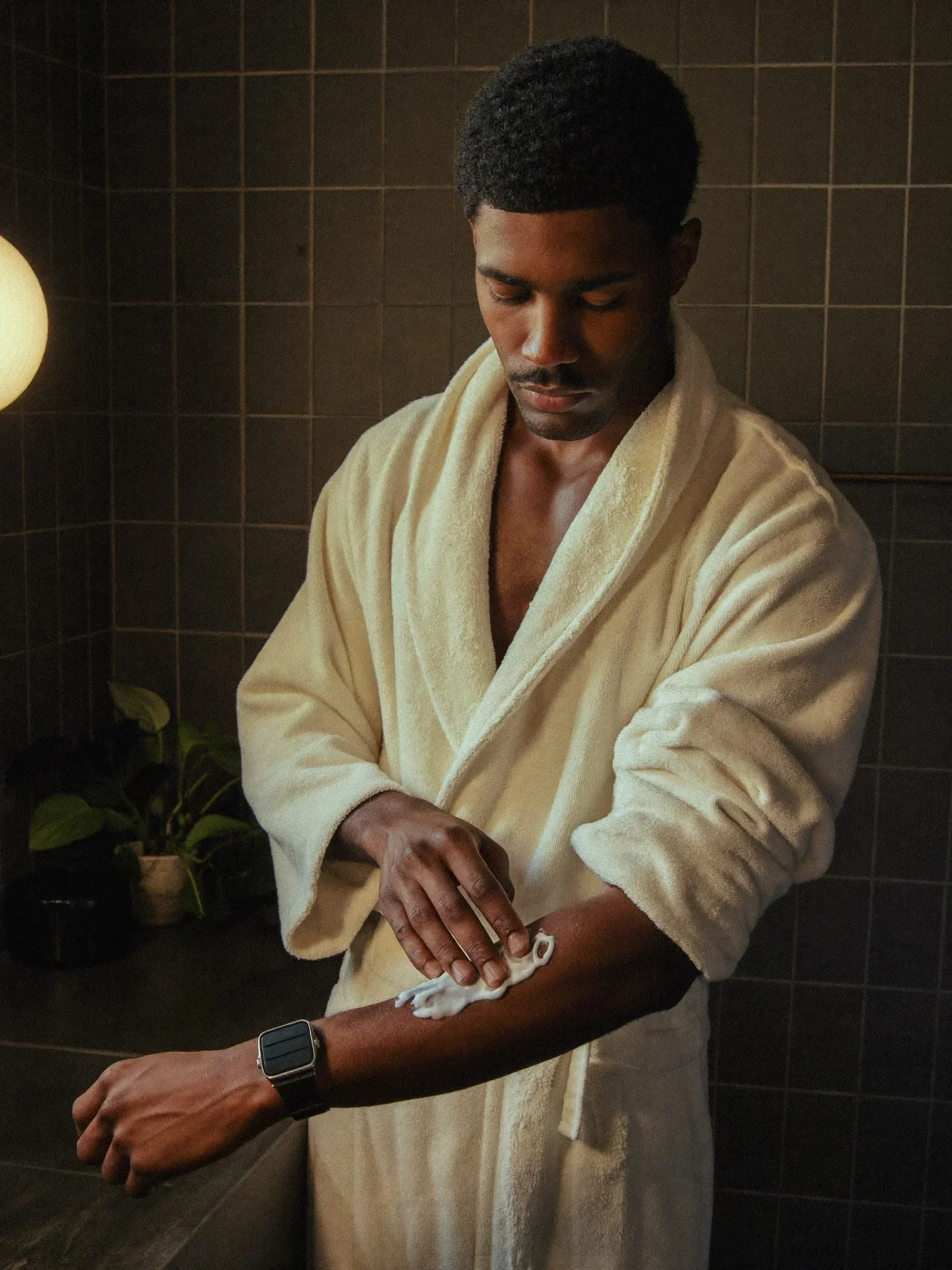 Black man in a cream robe applying body butter to his forearm