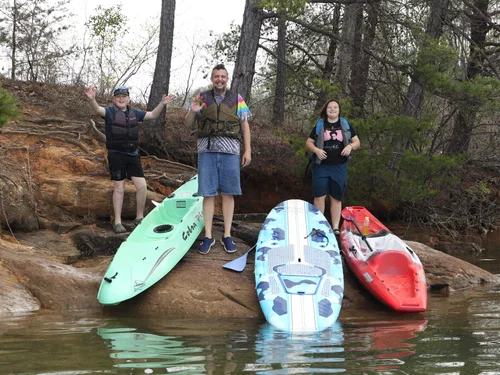 Rachel Stepek’s son Luke, husband Nathan, and child Red standing in life jackets with two kayaks and a paddleboard at a rocky wooded bank on Lake Keowee