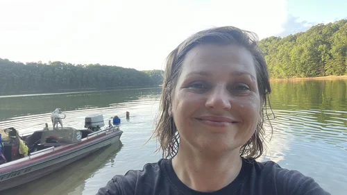 Rachel Stepek beside the family’s 1982 Ranger bass boat at Laurel River Lake, with her husband Nathan and son Luke swimming behind her and Piggy the dog standing on the boat