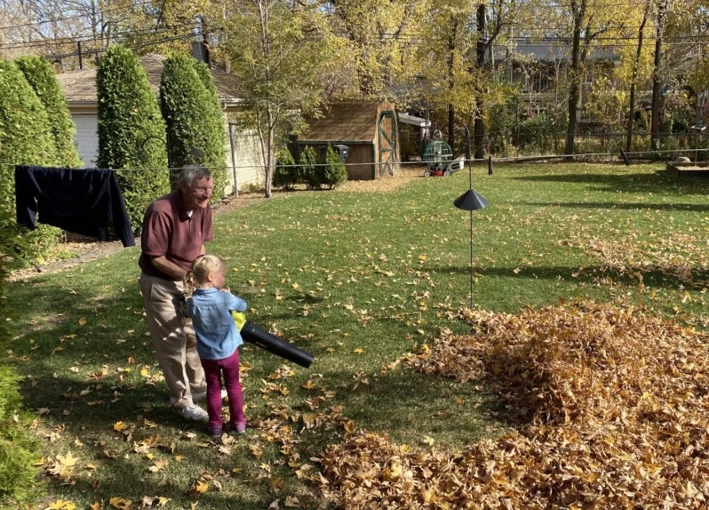 An older adult and a young child blowing leaves together in a sunny backyard