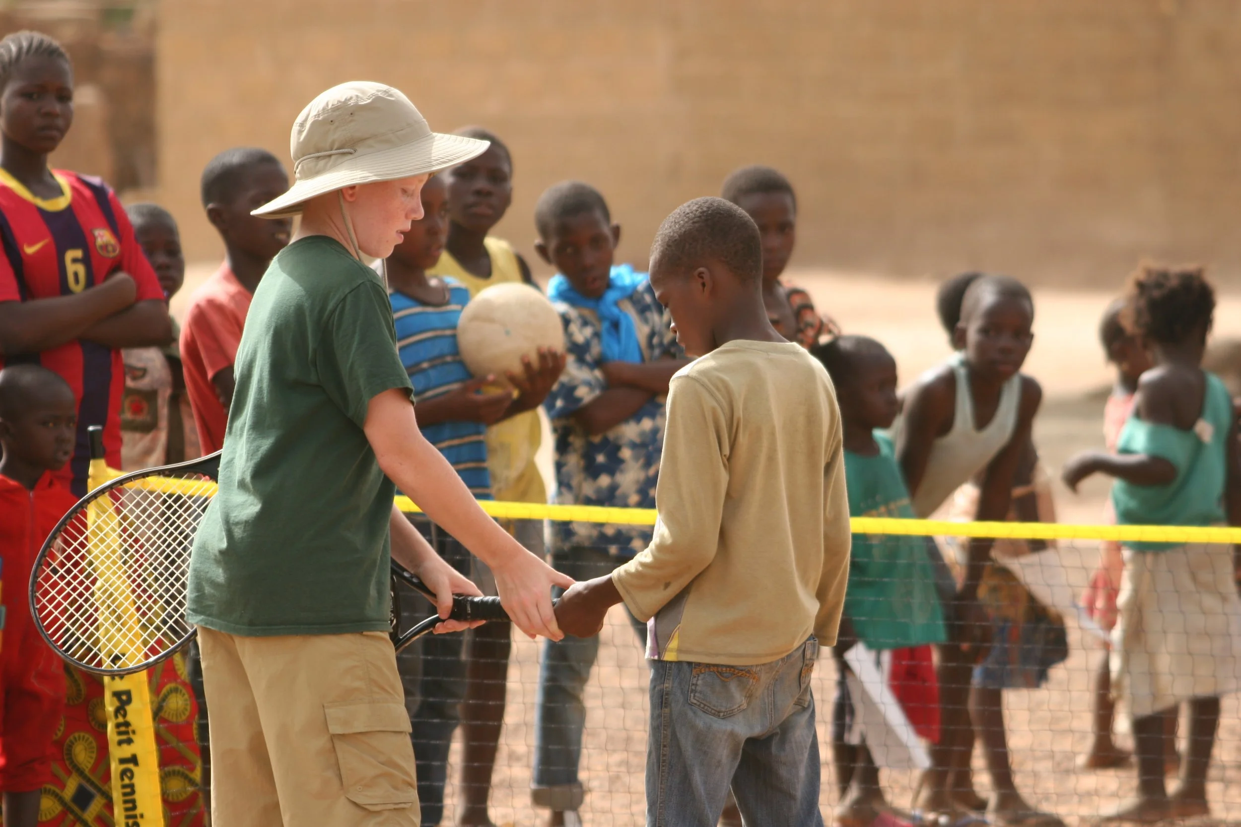 A young volunteer holding a racket beside a player at a tennis net in Mali