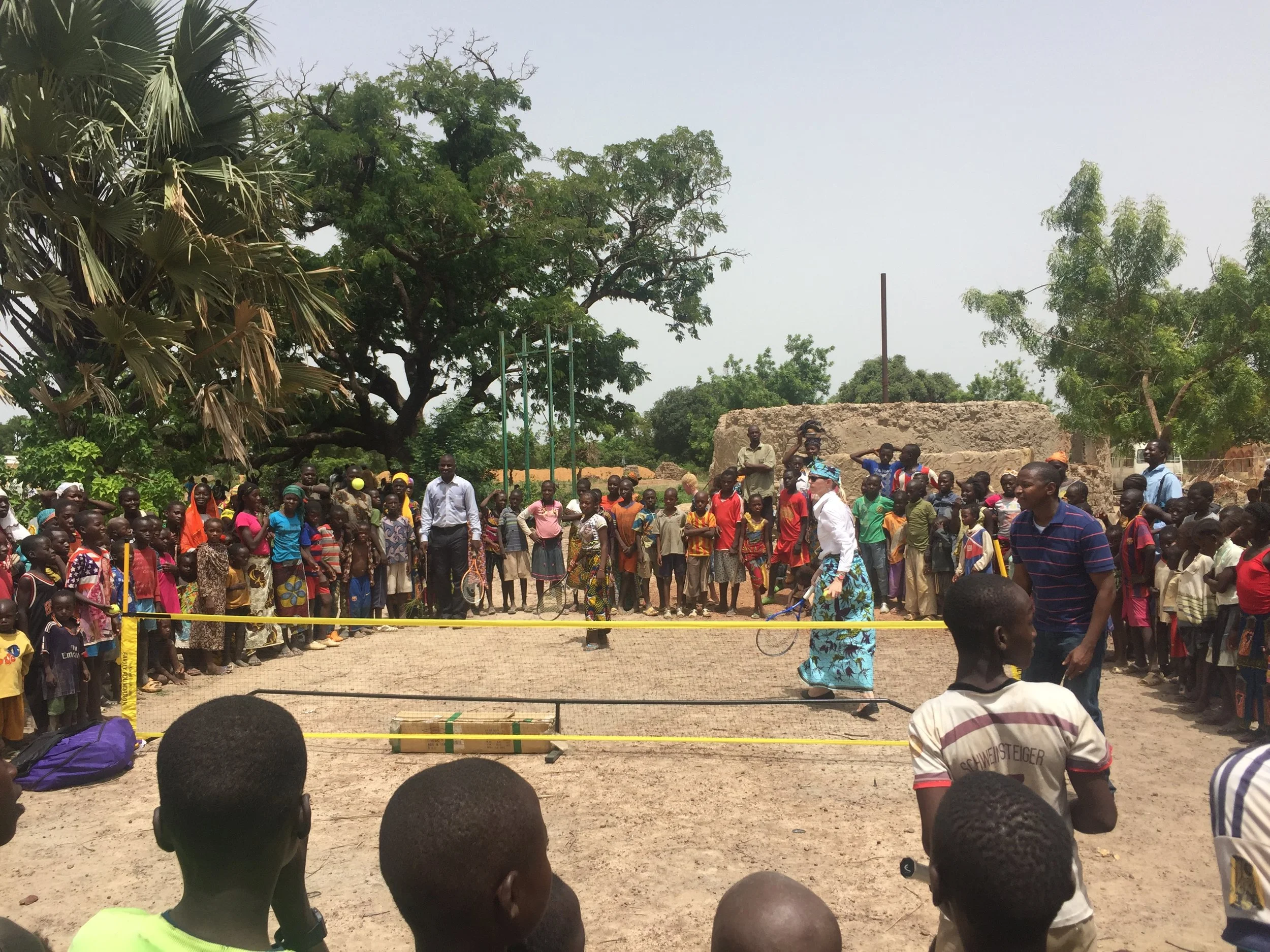Children and adults gathered around a tennis net on an earthen playing area in Mali