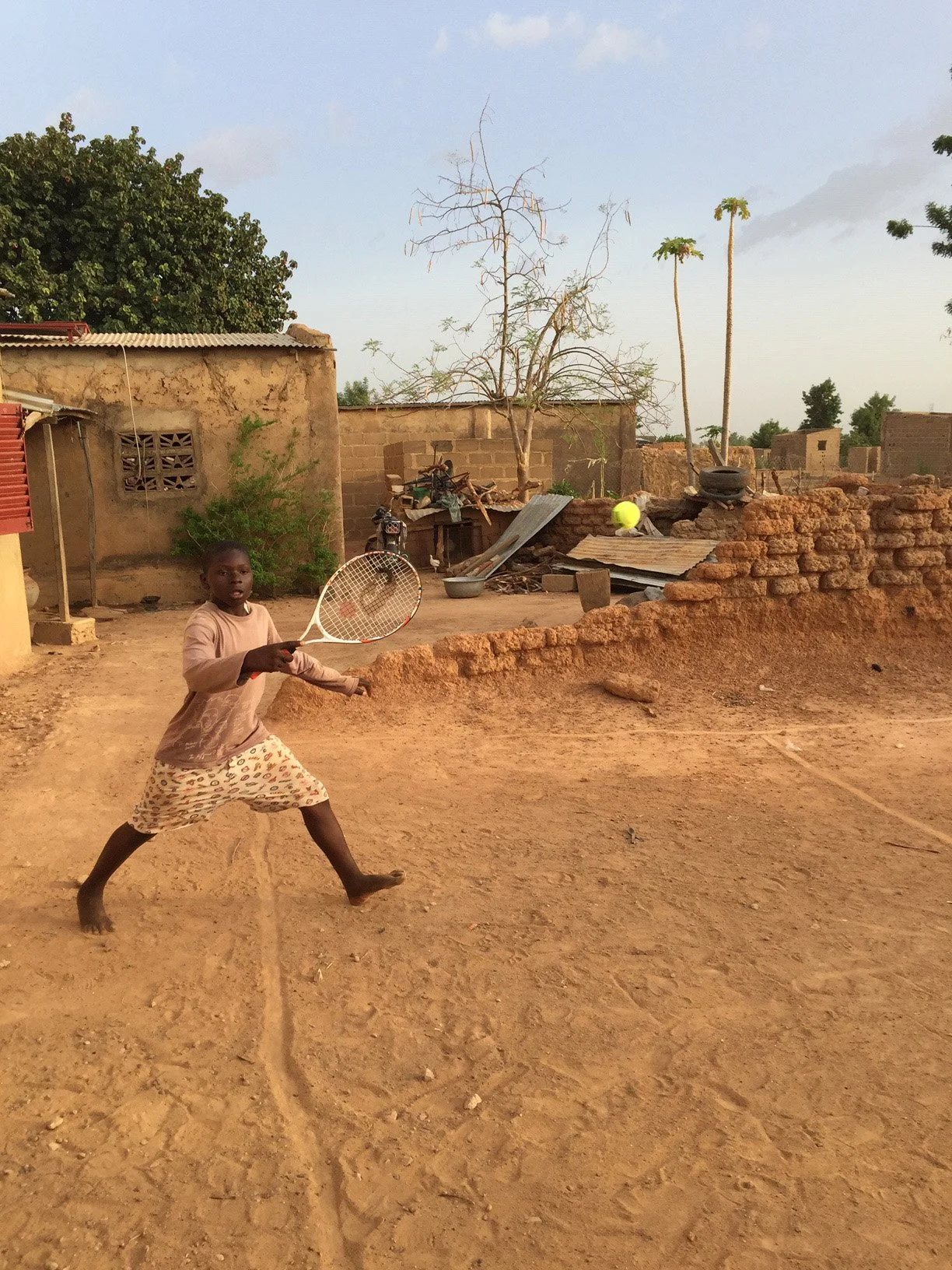 A young player in Mali striking a tennis ball on an earthen playing area, with village buildings and trees behind him