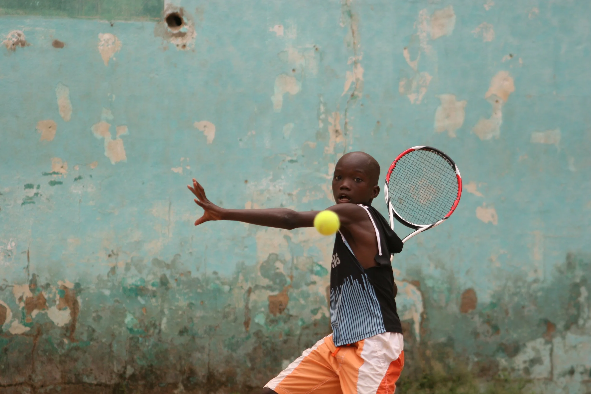 A young player in orange shorts preparing a tennis stroke against a blue wall in Mali