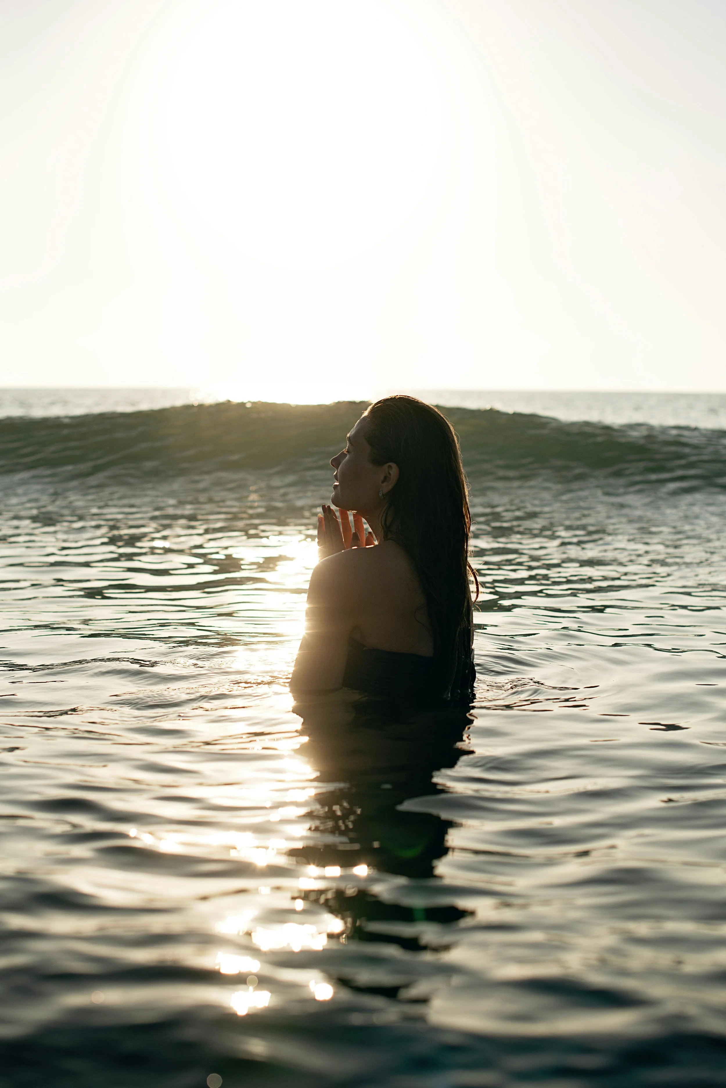 Woman standing in ocean at golden hour, hands in prayer