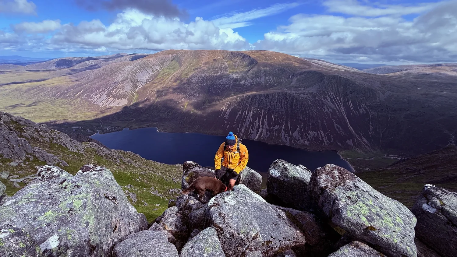 A person in an orange jacket pauses beside a Scottish loch, with a dog nearby and hills beyond.