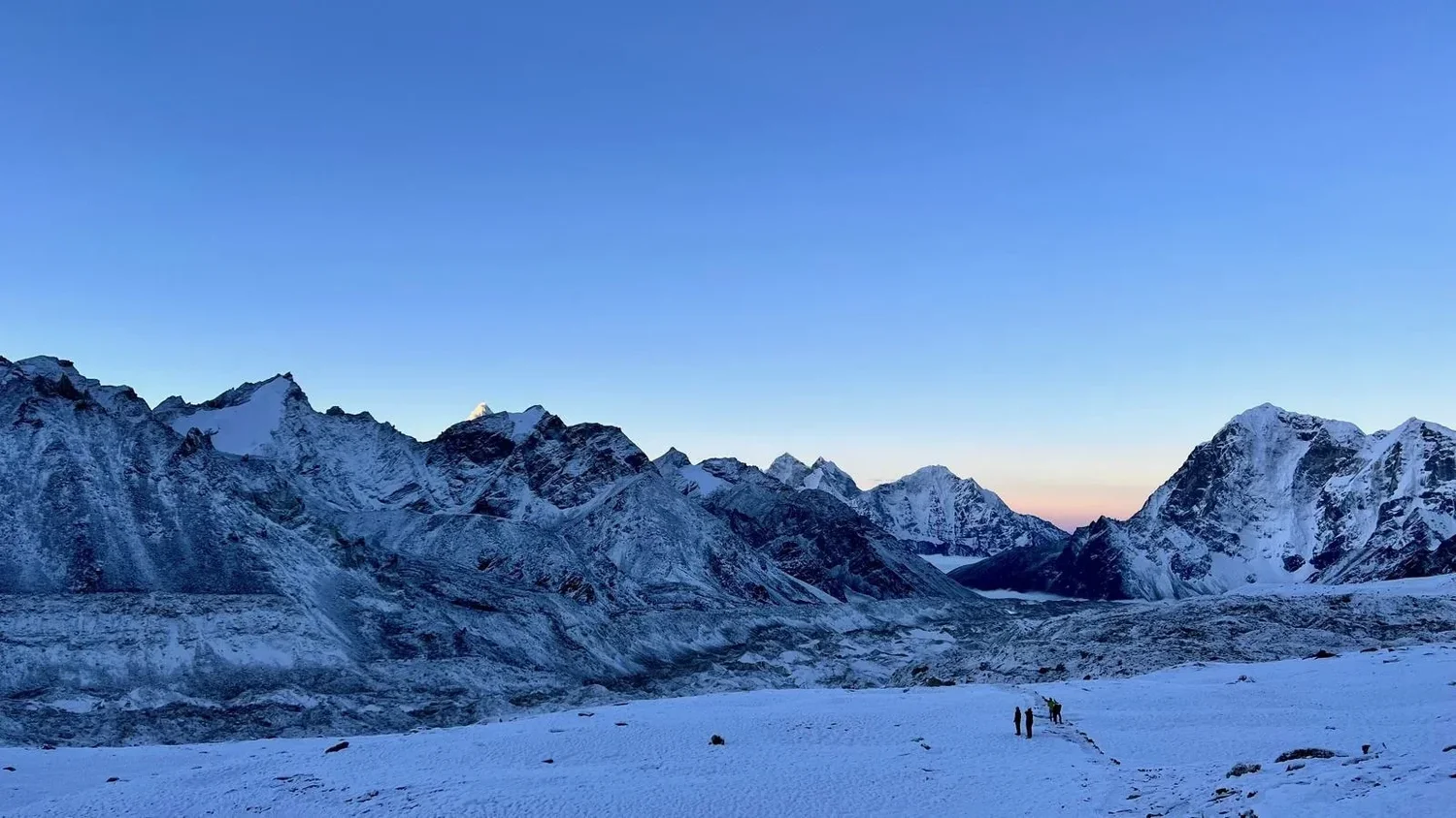 Himalayan peaks at dawn, with distant silhouettes following tracks across snow.
