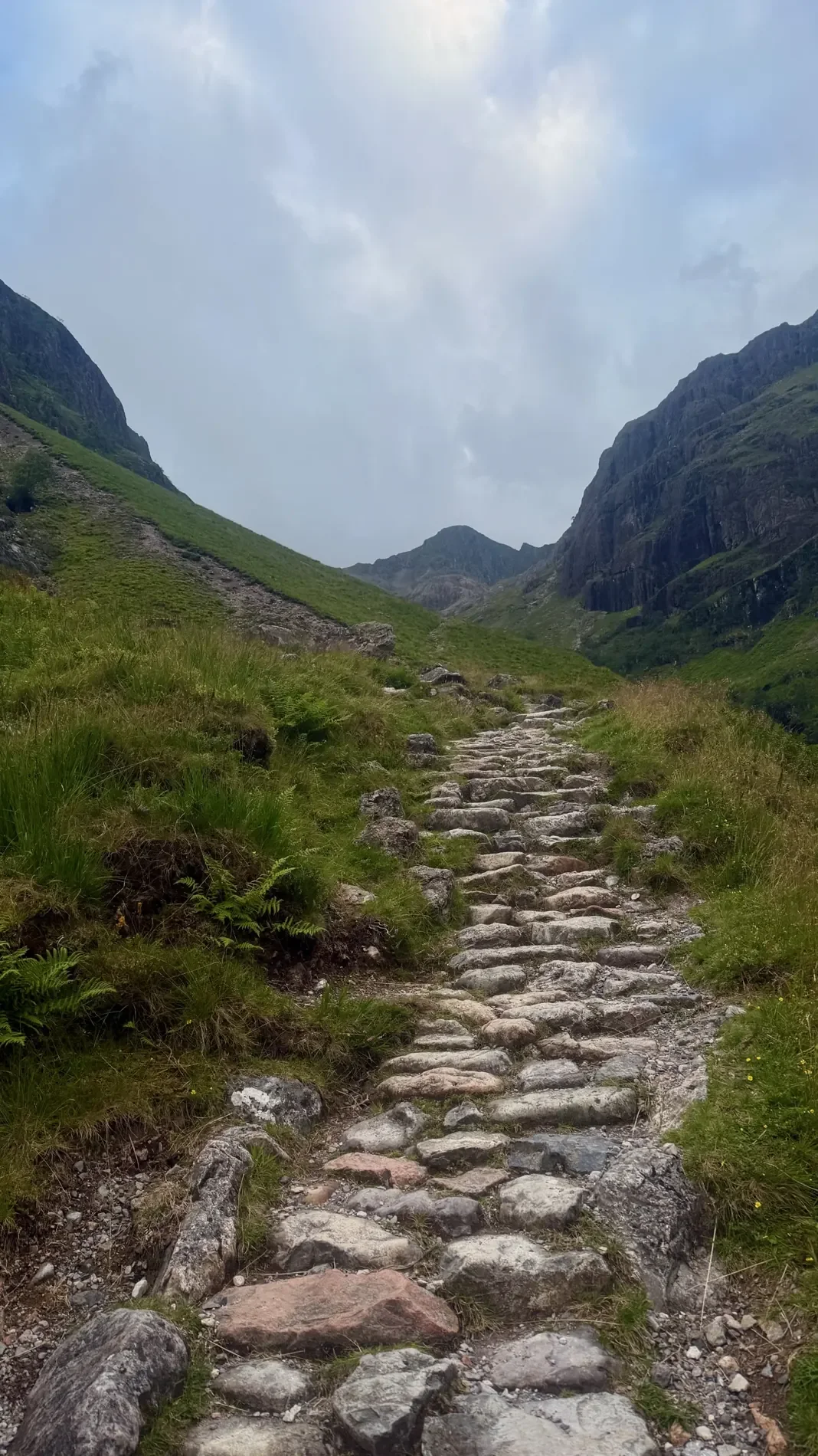 A stone path leads into a dramatic Scottish mountain glen beneath steep green slopes.