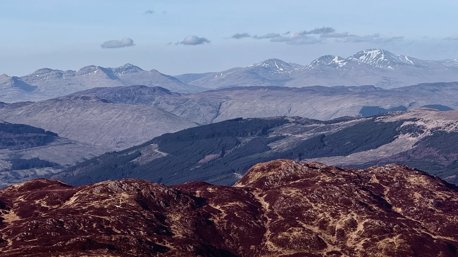 Heather-covered Scottish hills open towards distant mountains with patches of snow.
