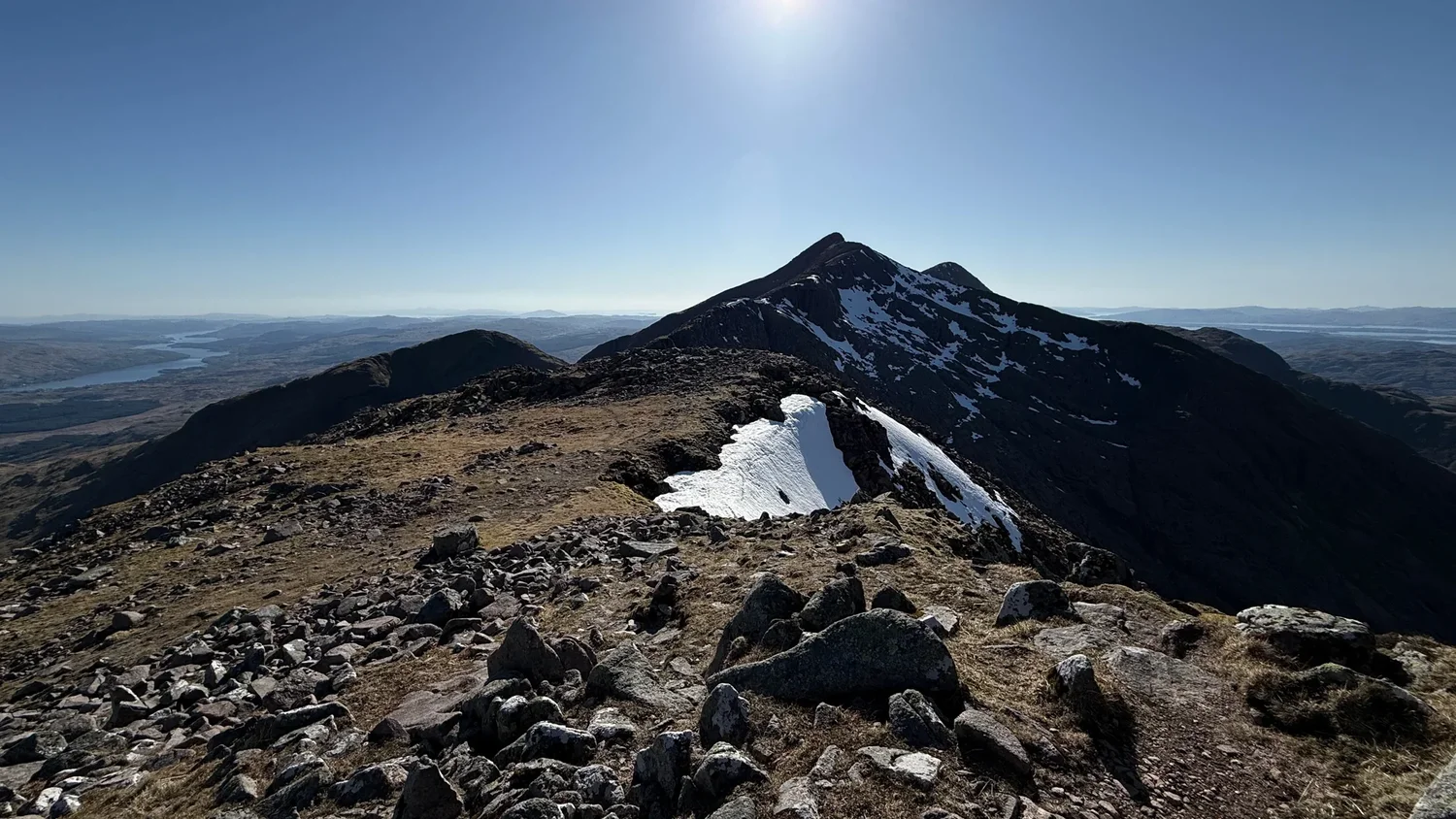 A rocky Scottish ridge leads towards a mountain summit, with patches of snow along its edge.