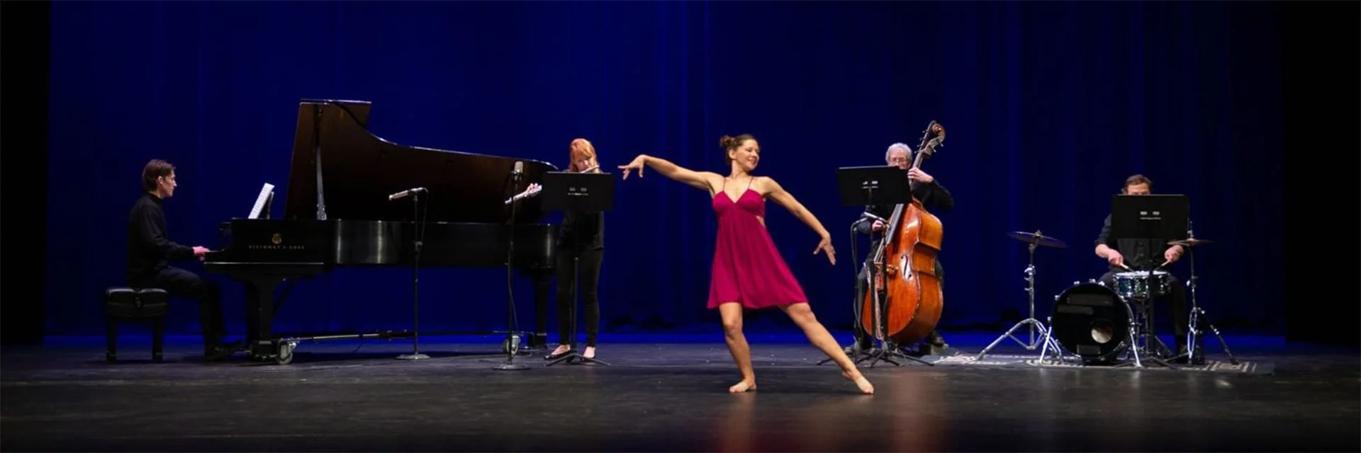 A TCM concert in performance — pianist, flutist, cellist, drummer, and dancer on stage against a deep blue curtain