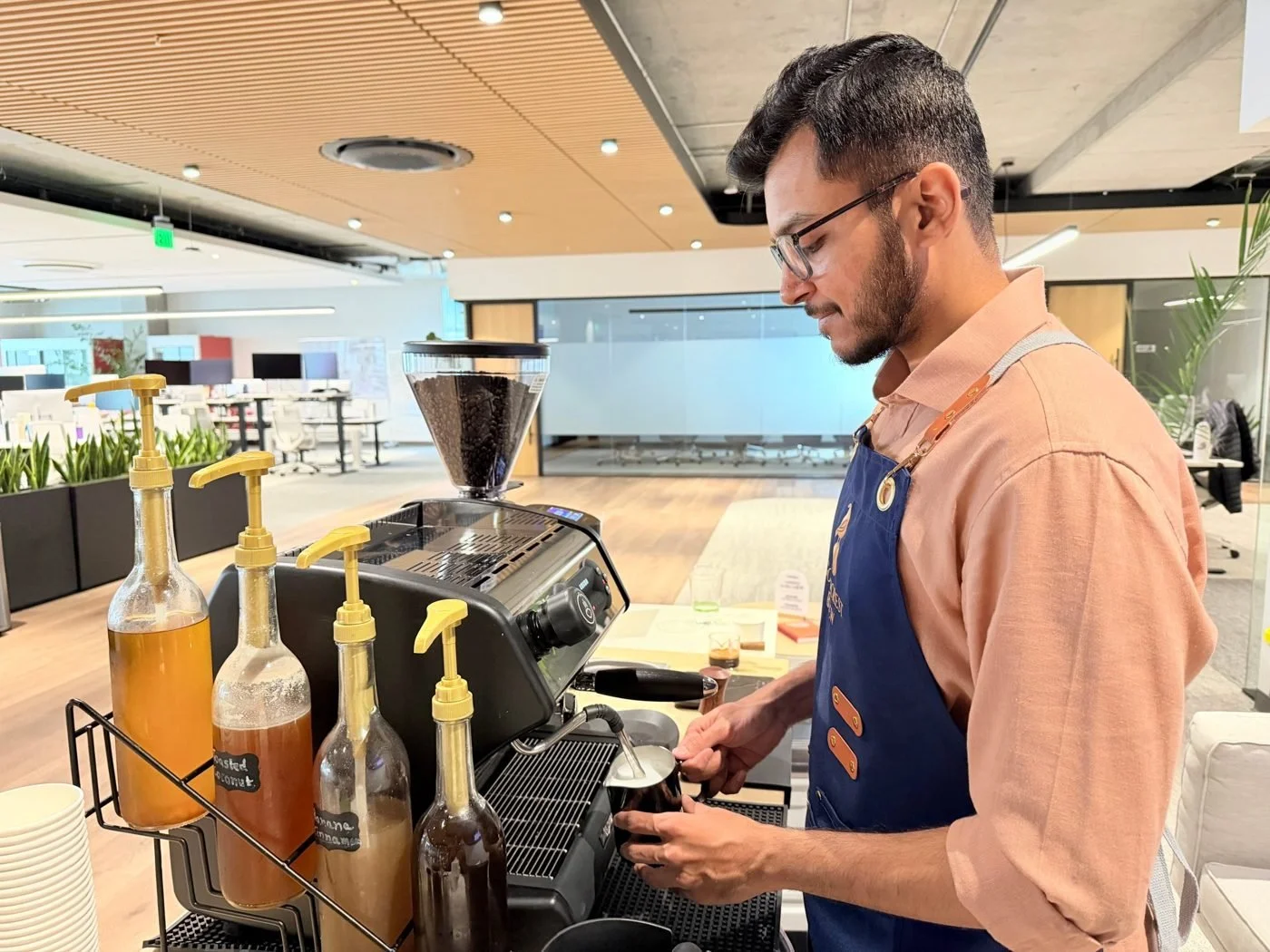 Barista steaming milk at a Goldcrest Brew espresso cart with house-made syrups in an open-plan office